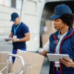 Black female worker using digital tablet while doing package inventory before the delivery. Her coworker is in the background.