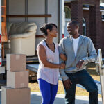 African American couple (30s) with moving truck in front of brick house, loading or unloading boxes and furniture.